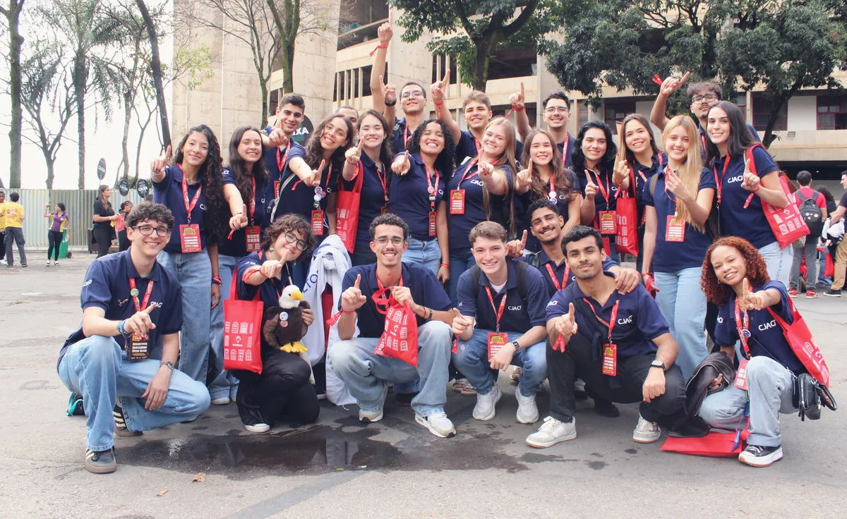 Cerca de trinta membros da CJA de polo azul-marinho e crachá do evento, agachados e em pé, apontam para a câmera e sorriem.