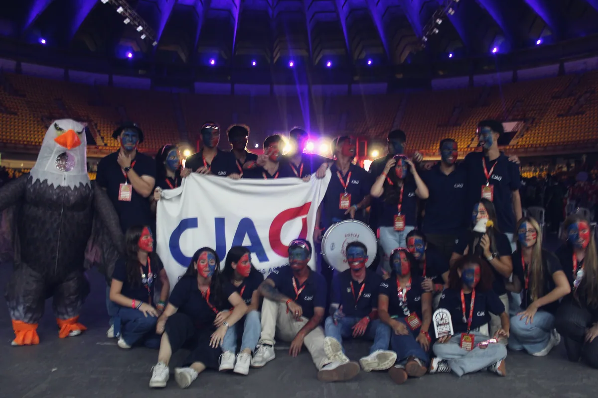 O time da CJA à noite, com a bandeira da CJA e a águia mascote, sob as luzes roxas da arena.