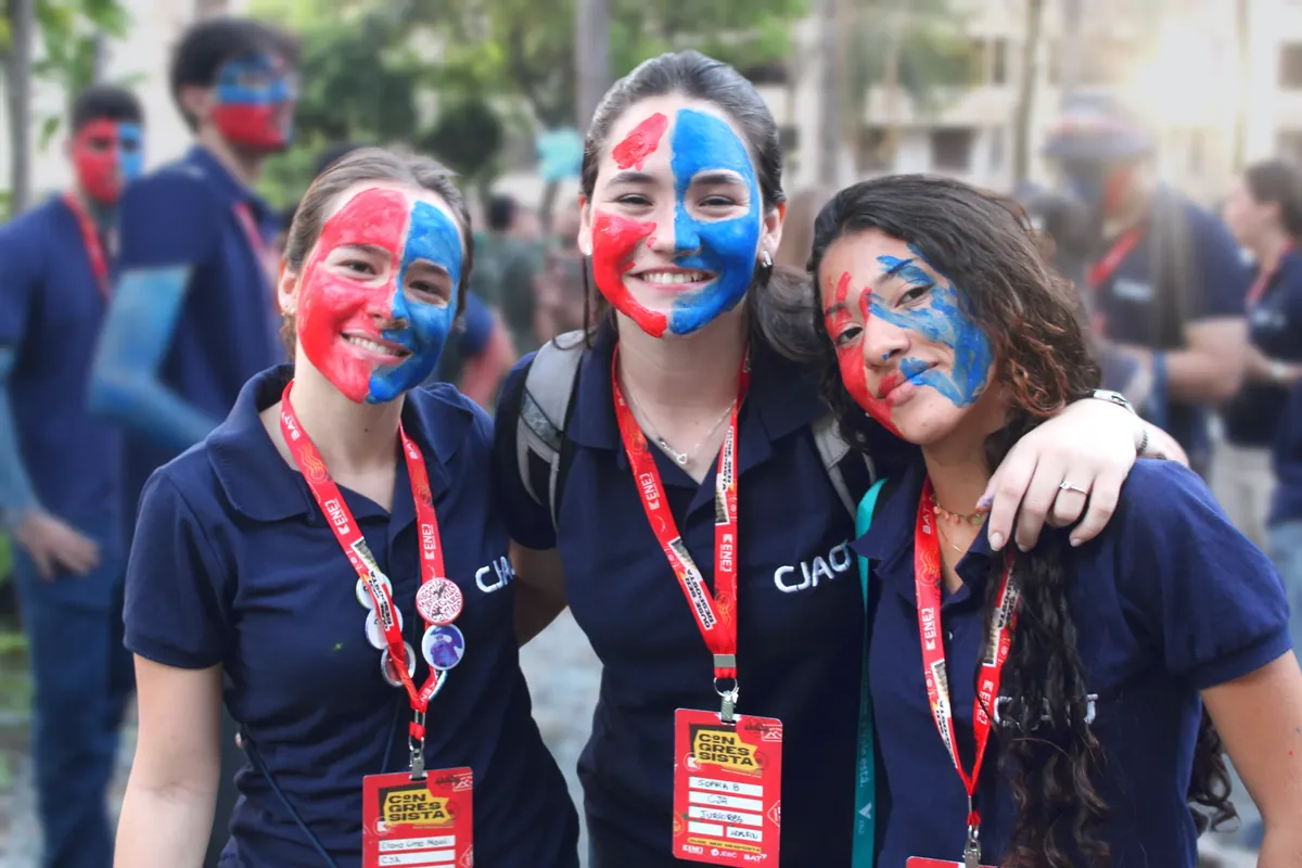 Três membros da CJA abraçados no ENEJ 2026, com o rosto pintado metade de vermelho e metade de azul.