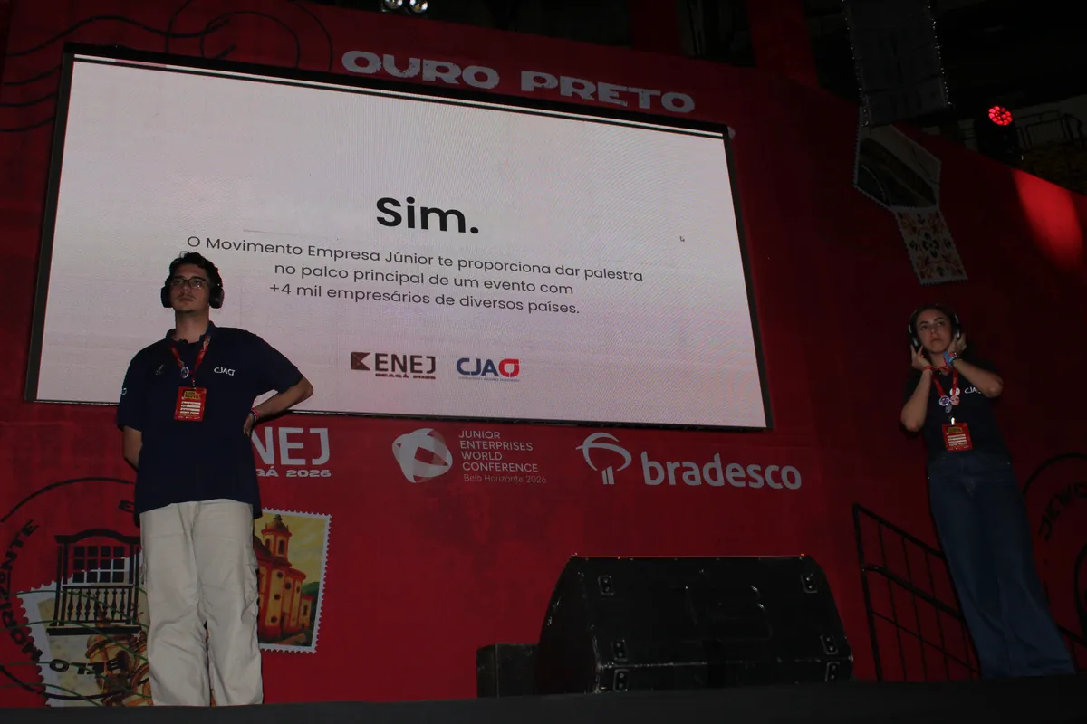 Dois membros da CJA no palco principal do ENEJ 2026, diante do telão com a apresentação da CJA.