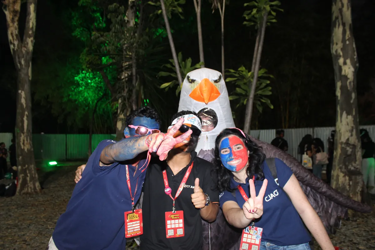 Três membros da CJA com o rosto pintado de vermelho e azul fazem pose à noite ao lado da águia mascote, no ENEJ 2026.