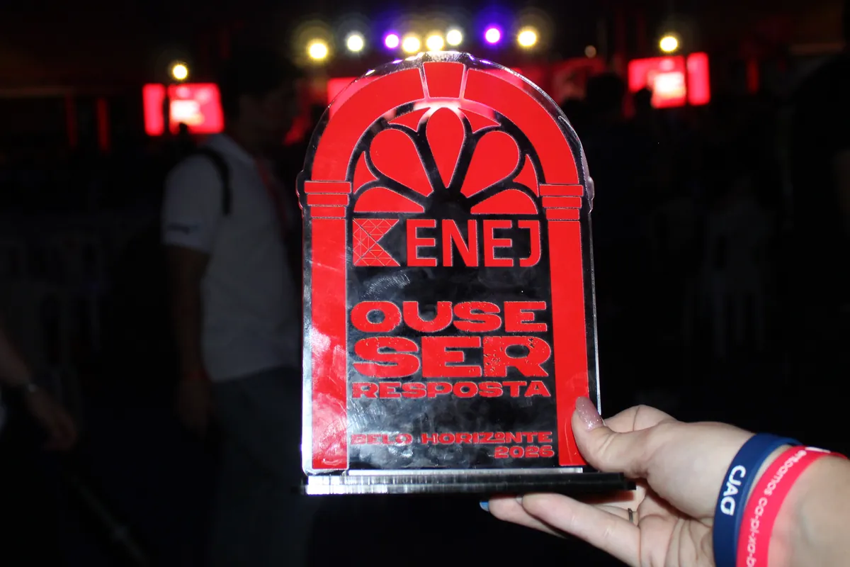 Uma mão segura o troféu vermelho do ENEJ 2026, com a arena iluminada ao fundo.